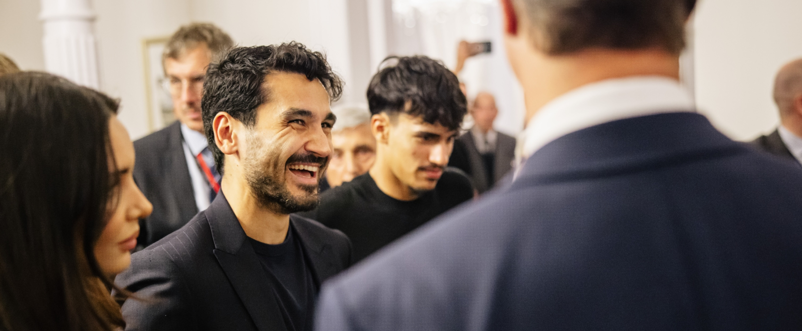 Ministerpräsident Wüst steht mit dem Rücken zum Betrachter, er spricht mit Ilkay Gündogan
