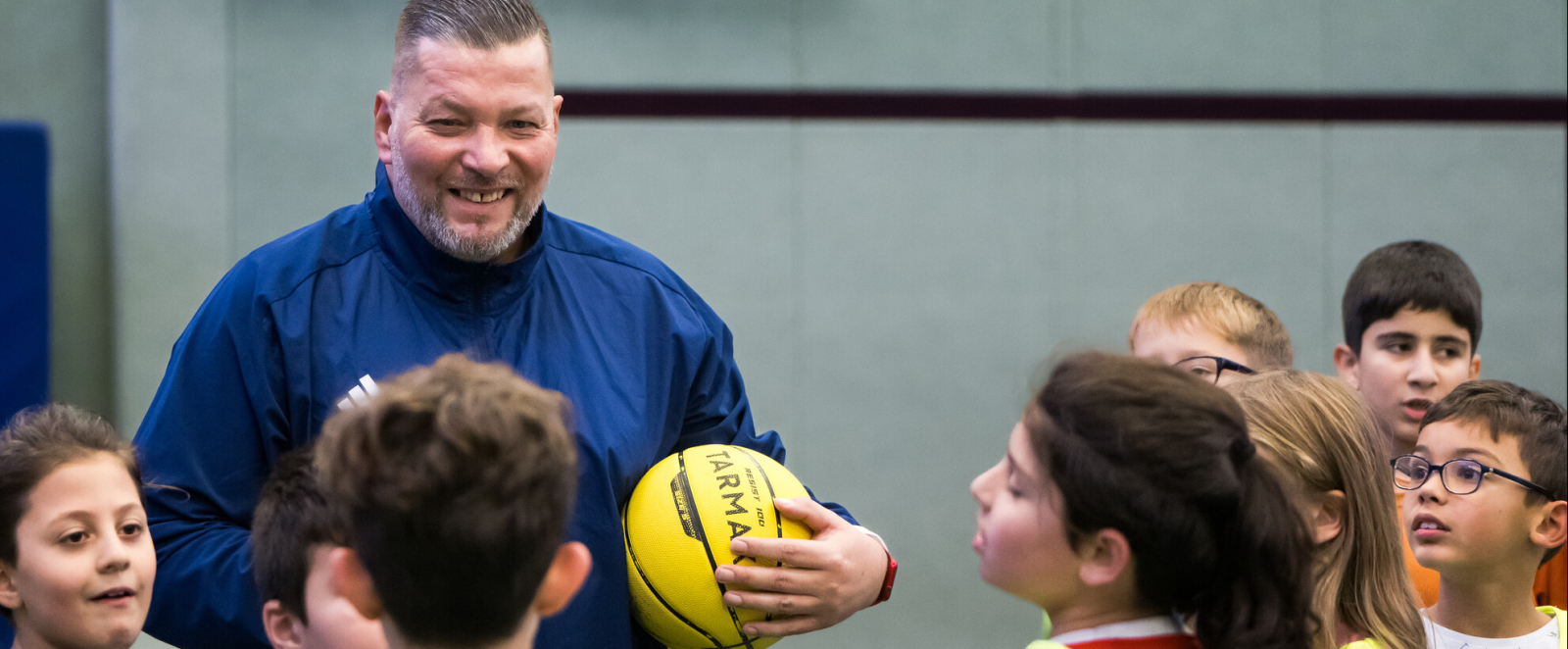 Mann in blauem Trikot hält einen gelben Ball und steht vor einer Gruppe Kinder