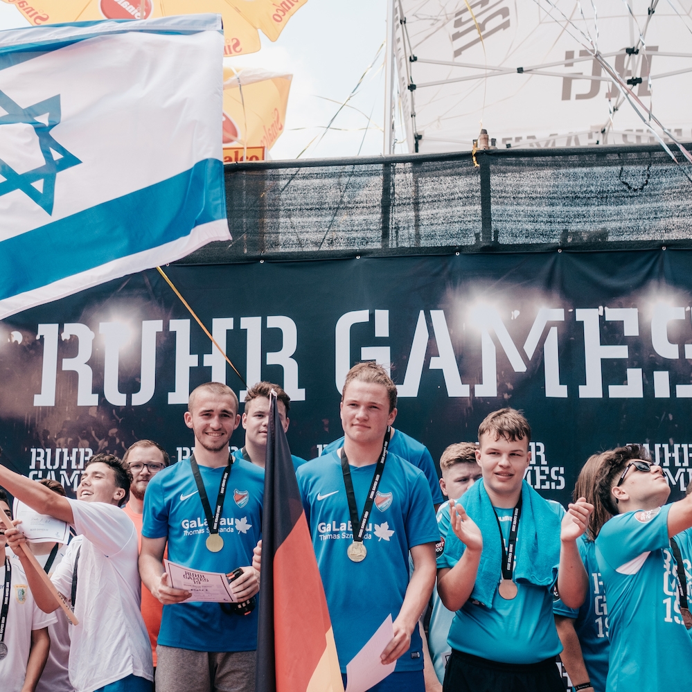 Viele Sportler stehen auf einer Bühne, einige Sportler halten Flaggen. Man sieht die Flaggen von Deutschland und Israel. Auf der Rückwand steht weiß auf schwarzem Grund "Ruhr Games".