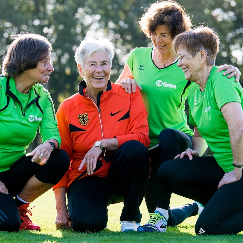 Vier ältere Damen bei einer Teambesprechung beim Sport. Drei Damen haben ein grünes Oberteil an, eine Dame in der Mitte hat ein rotes Oberteil an. Alle haben schwarze Sporthosen an.