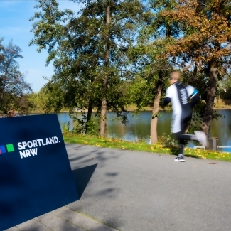 Jogger läuft an einem See an einem Sportland.NRW Schild vorbei