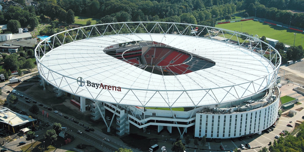 Stadion BayArena  Leverkusen aus der Vogelperspektive. Ein offenes Stadion mit überdachten Tribühnen im kreisdunden Design, Dach und Außenfassde in weiß, oben außen herum eine Stahlring-Konstruktion mit Verstrebungen.