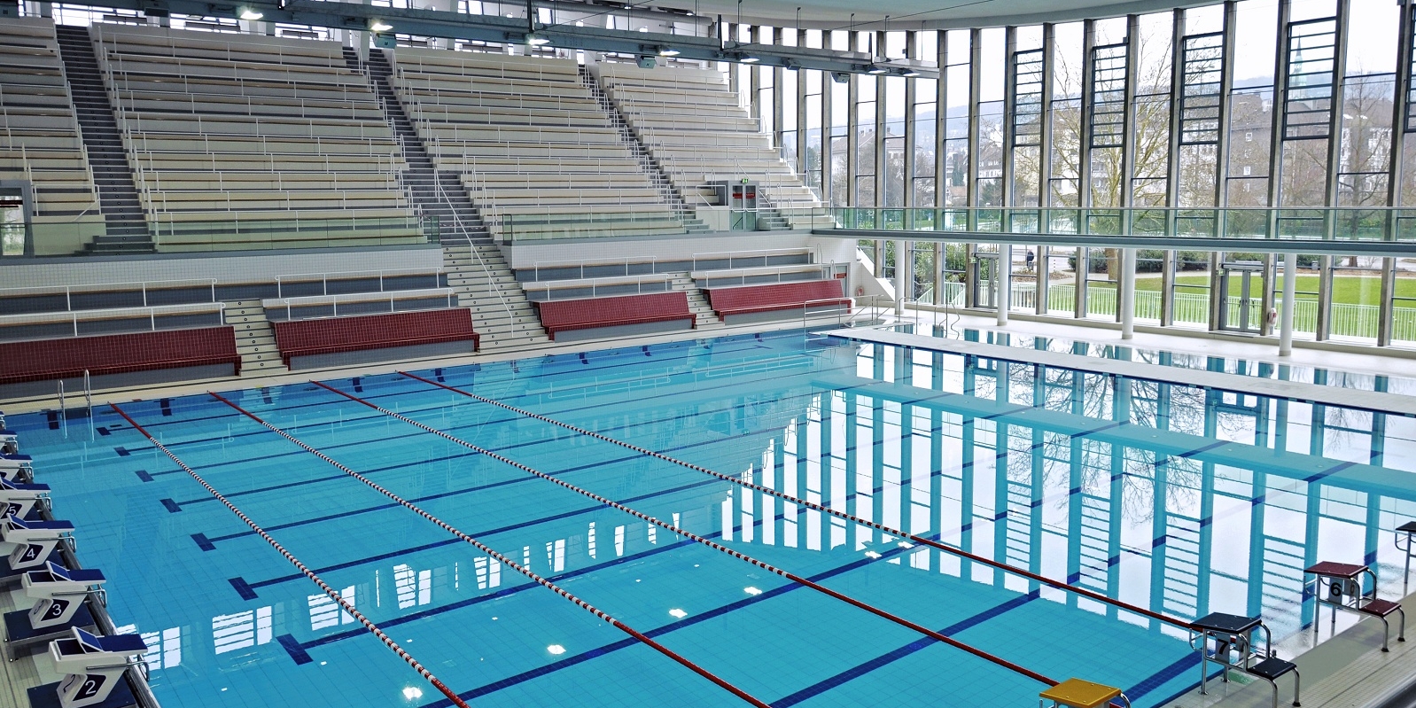 Große Schwimmhalle mit Sprungböcken, sowie eine große leere Tribüne für Zuschauer.