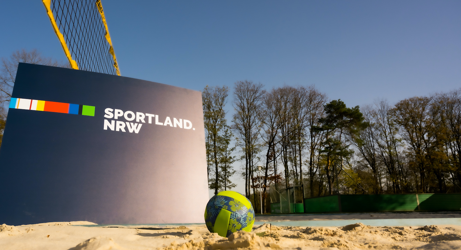 blaues Schild mit Aufschrift "Sportland.NRW" vor einem Beachvolleyballnetz, ein Ball liegt im Sand davor
