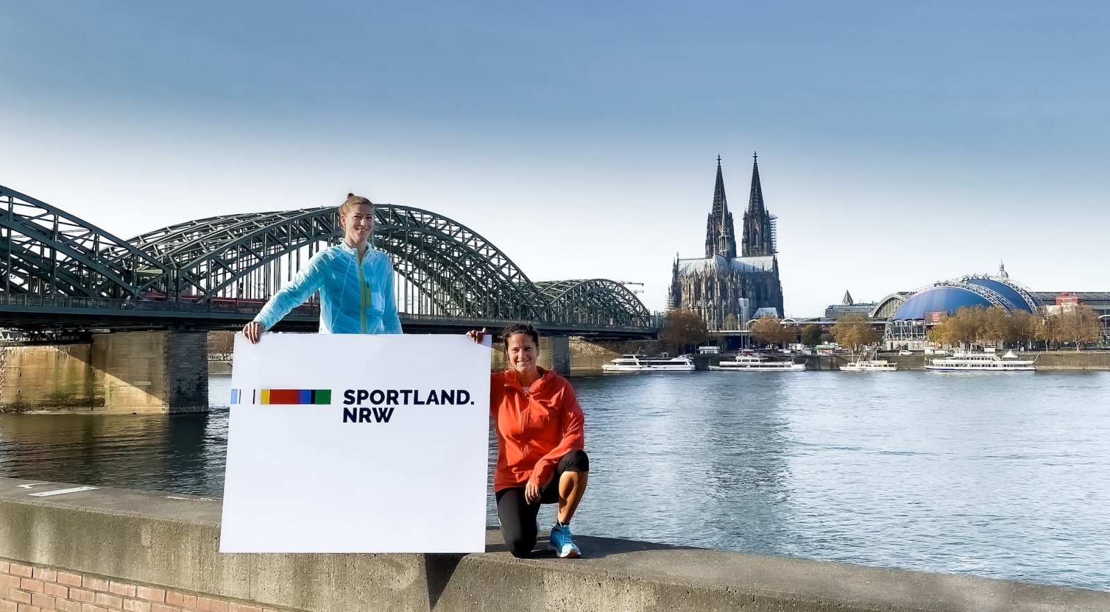 Vor dem Kölner Dom: Zwei Sportlerinnen halten den Sportland.NRW-Banner .
