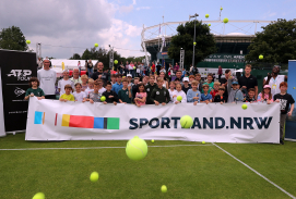 Auf einem Rasen-Tenniscourt bei den Terra Wortmann Open sind Kinder hinter einem Sportland.NRW-Banner und werfen gelbe Bälle