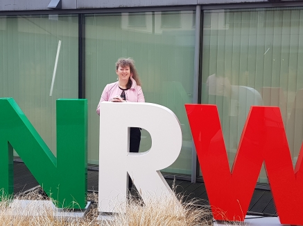 Staatssekretärin Andrea Milz steht vor einer Fensterfront, vor ihr die drei Buschtaben N R W in grün, weiß und rot.