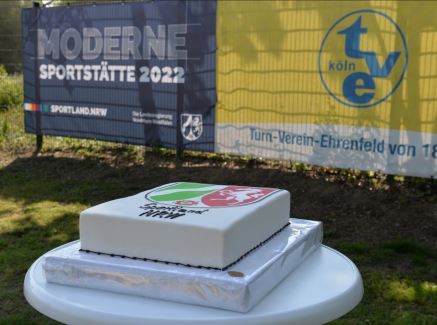 Auf einem Rasen steht ein weißer, runder Stehtisch, darauf ein Kuchen mit NRW-Wappen und dem Schriftzug Sportland.NRW.