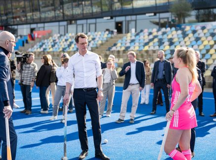 Ministerpräsident Wüst mit Hockeyschläger neben einer Hockeyspielerin in pinkem Dress im Stadion