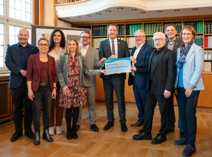 Gruppe von Personen in einem Raum mit Holzboden, ein Mann in der Mitte hält einen Bescheid in der Hand