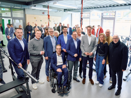 Hendrik Wüst umgeben von mehreren Personen, davon einer im Rollstuhl, in einer Halle mit Fenstern im Hintergrund