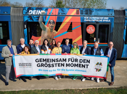 Andrea Milz und weitere Personen mit einem Banner der KRR-Bewerbung vor einer Straßenbahn im "Olympia-Look"