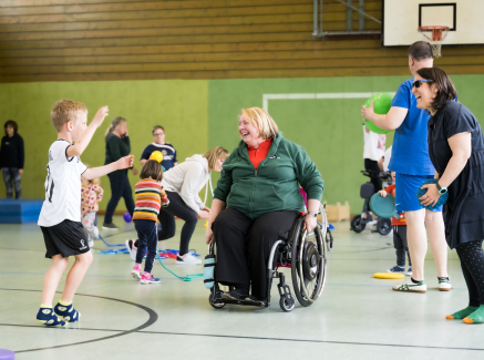 Ältere Frau in Sportkleidung im Rollstuhl ist umgeben von jüngeren Sportlern in eienr Sporthalle