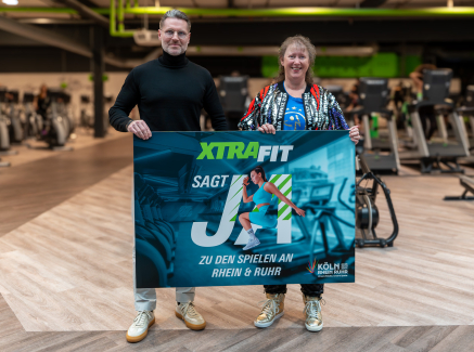 Andrea Milz neben einem Mann in einem Fitnessstudio, beide halten ein Banner zur KRR-Kampagne