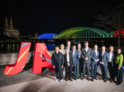 Eine Gruppe von Menschen steht vor großen bunten Buchstaben, im Hintergrund sind eine beleuchtete Brücke und die nächtliche Skyline zu sehen.
