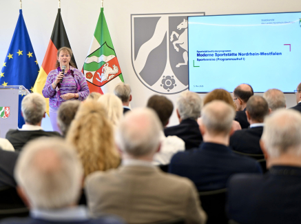 Andrea Milz vor drei Fahnen und Videobildschirm, sie spricht zu Teilnehmenden einer Pressekonferenz