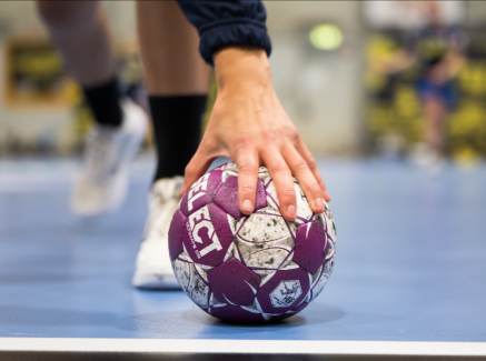 Handball auf einem blauen Boden, im Hintergrund sind Hände zu sehen