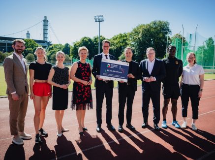 Ministerpräsident Wüst umgeben acht Personen auf einer Tartanbahn in einem Stadion, er hält ein Schild in der hand