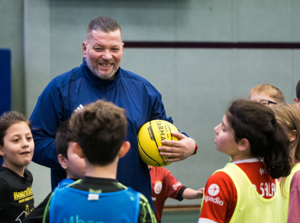 Mann in blauer Sportkleidung hält einen gelben Ball, er ist umringt von Jungen und Mädchen 