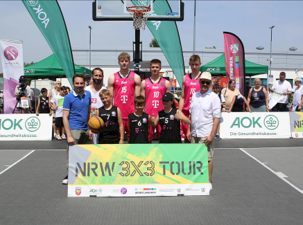 Gruppenfoto beim Basketball-Event „NRW 3x3 Tour“ mit Spielern in rosa-schwarzen Trikots, im Hintergrund sind Banner und Zuschauer zu sehen.