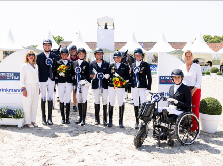 Team von Para Dressurreiterinnen und -reitern, eine davon in einem Rollstuhl, vor einem Hindernisparcours