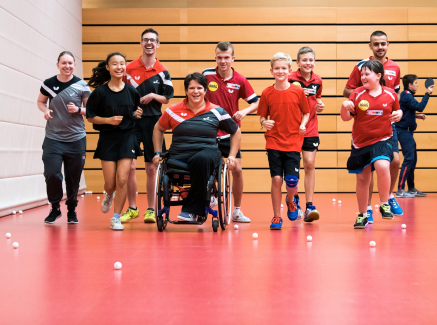 Gruppe von Sportlerinnen und Sportlern in einer Halle mit rotem Boden, eine davon in eiem Rollstuhl