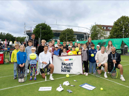 Gruppe von Kindern halten ein Banner mit "Sportland.NRW" auf einem Rasen-Tennisplatz