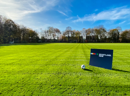 Grüne Rasenfläche, umgeben von Bäumen, auf der ein Sportland.NRW-Schild steht, danaeben liegt ein Fußballball