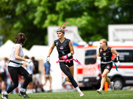 Junge Frau in schwarzer Sportkleidung spielt Flag Football auf einer Wiese, im Hintergrund stehen Fahrzeuge