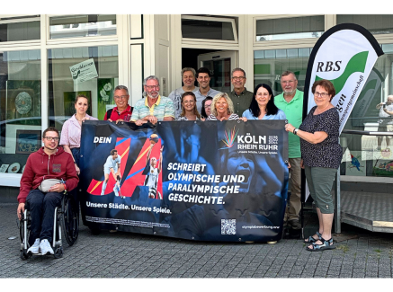 Gruppe von Menschen, davon einer in einem Rollstuhl, vor einem Gebäude. Sie halten ein Banner der Bewerbung KölnRheinRuhr