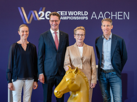 Ministerpräsident Wüst und drei Pferdesport-Athletinnen und -Athleten vor einem Banner der Reit-WM in Aachen, vor ihnen ein goldfarbenes Schachfigur-Pferd