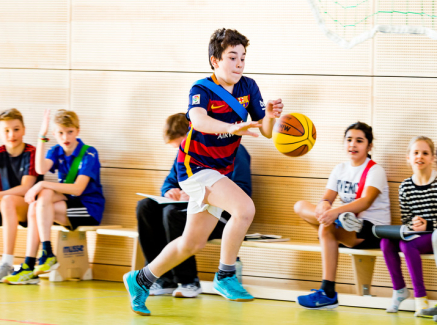 Schulikinder auf einer Bank in einer Turnhalle, davor ein Junge mit einem Basketball