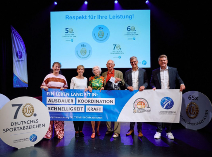 Sechs Personen halten einen Banner bei einer Veranstaltung, wo der 100-jährige Johann Otte als ältester Teilnehmer beim Deutschen Sportabzeichen ausgezeichnet wurde.