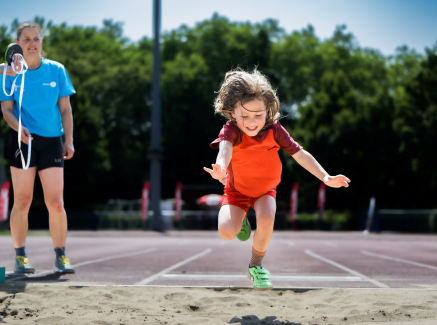 Junge Sportlerin springt in eine Sandgrube, links neben ihr steht eine Trainerin in blauem Shirt