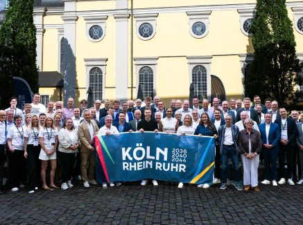Gruppe von Menschen vor einem beigefarbenen Altbau mit einem Banner der Bewerbung KRR