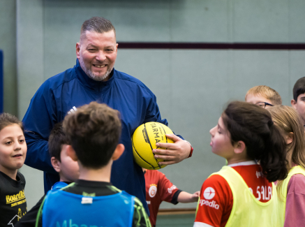 Mann mit blauem Triikot hält einen gelben Ball,, vor ihm Kinder in einem Kreis