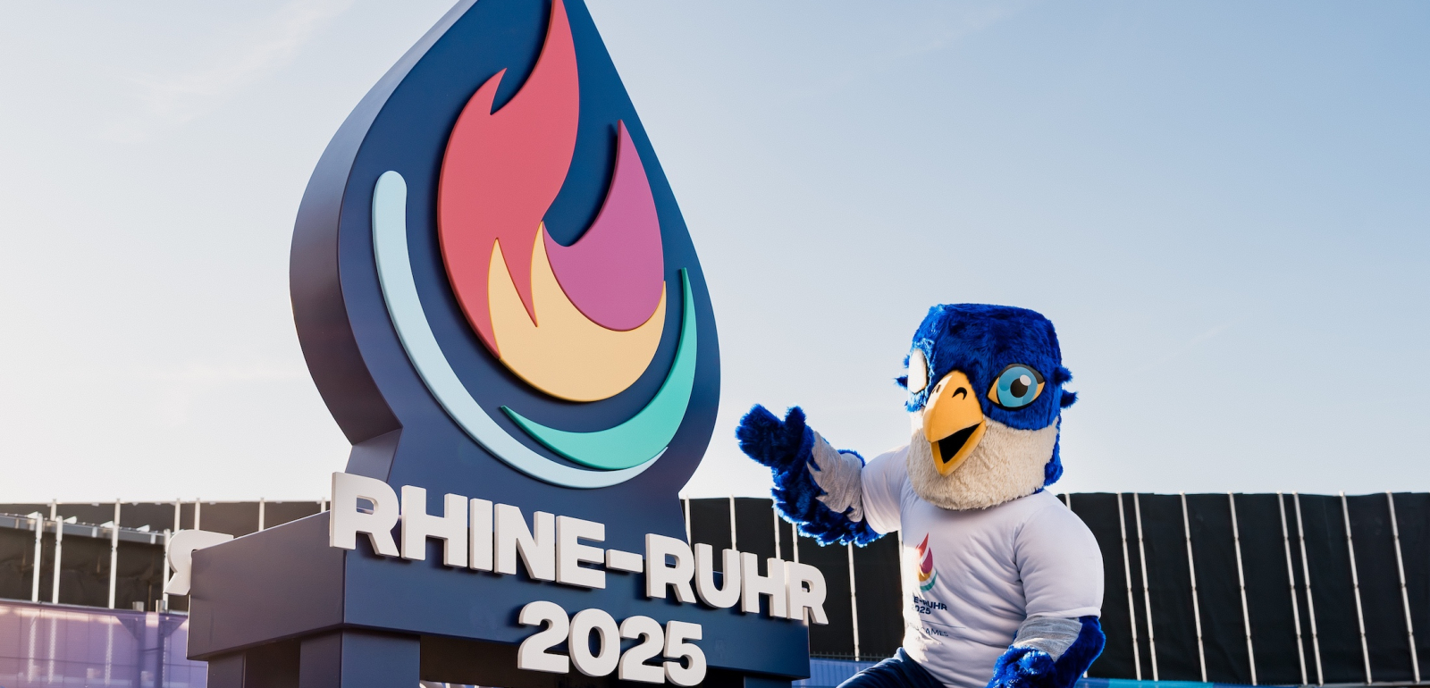 Das Maskottchen der Spiele vor dem Logo der Spiele