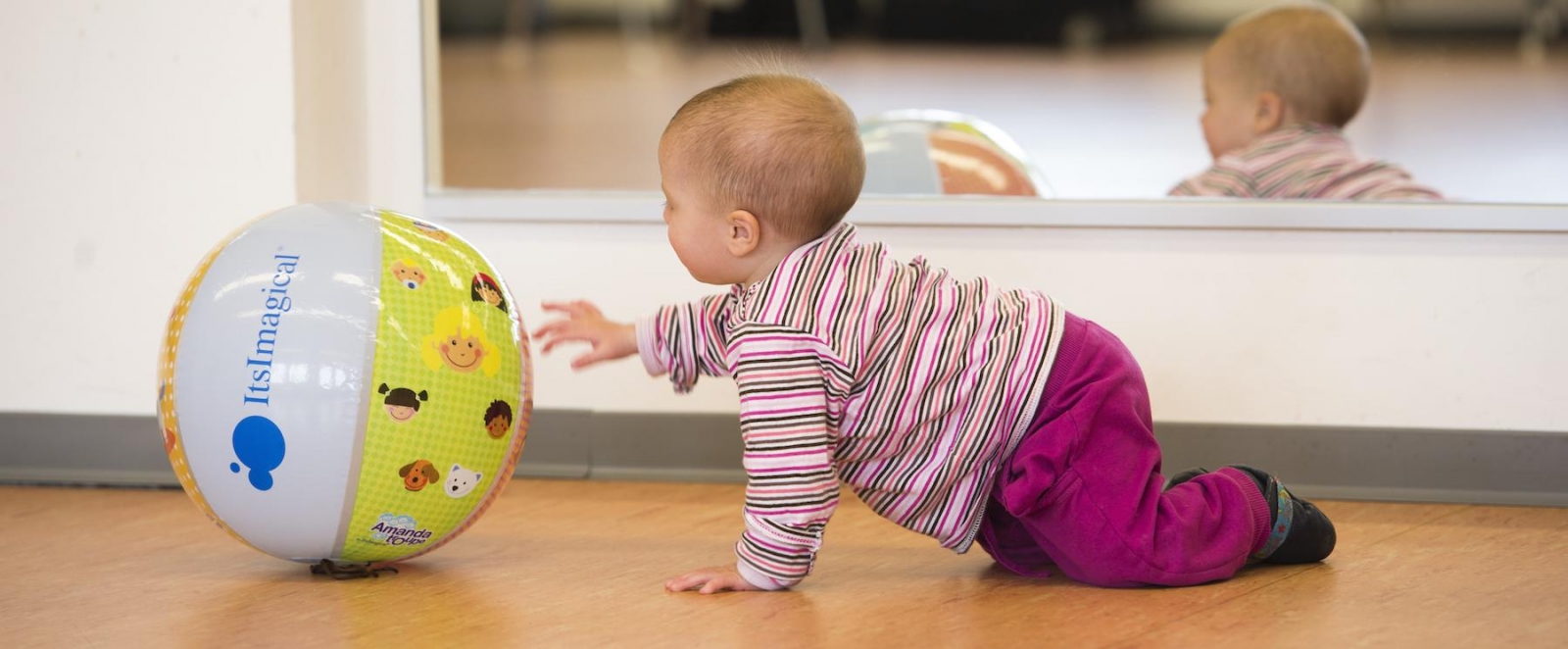 Ein Baby krabbelt über den Boden und streckt seine linke Hand nach einen bunten Plastikball aus.