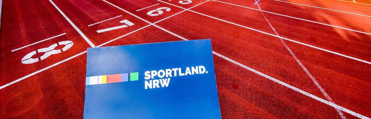 Ein blaues Schild mit dem Sportland.NRW-Logo liegt auf einer roten Tartanbahn.