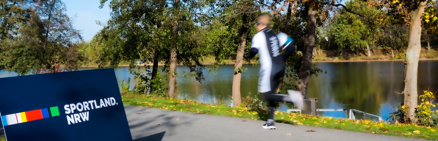 Blaues Sportland.NRW-Schild mit einem vorbeilaufenden Jogger