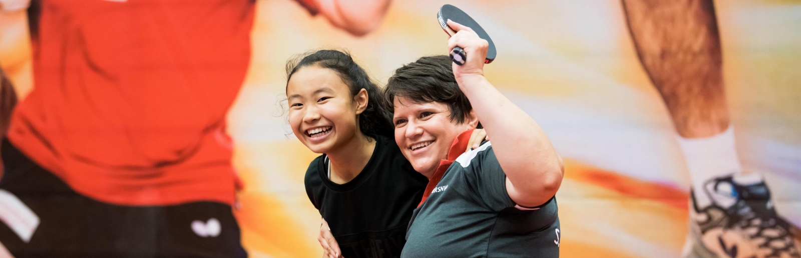 Zwei Tischtennisspielerinnen freuen sich, eine von ihnen sitzt im Rollstuhl. Im Hintergrund an der Wand ein großes Bild von einem Tischtennisspieler im roten T-Shirt und schwarzer, kurzer Hose.