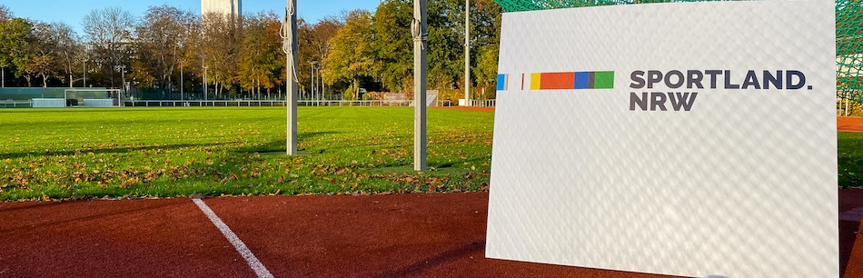 Ein weißes Schild mit dem Sportland.NRW-Logo steht auf einem Tartanbahn eines Sportplatzes, dahinter hängt ein grünes Netz.