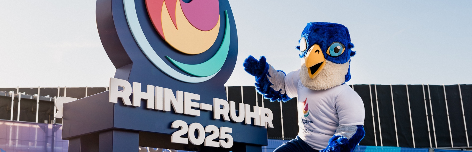 Das Maskottchen der Spiele vor dem Logo der Spiele
