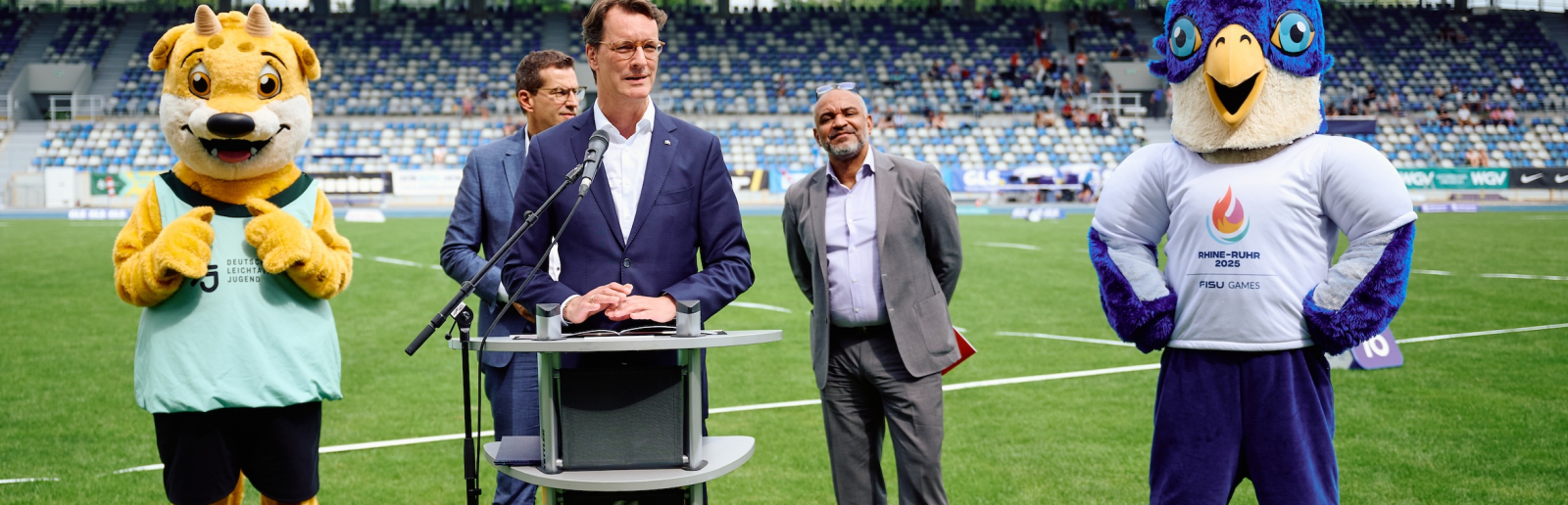 Hendrik Wüst auf dem Rasen im Lohrheidestadion mit Maskottchen und zwei Personen