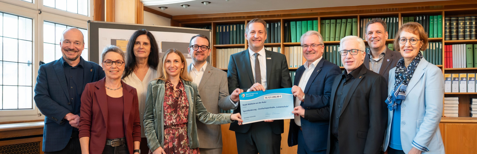 Gruppe von Personen in einem Raum mit Holzboden, ein Mann in der Mitte hält einen Bescheid in der Hand
