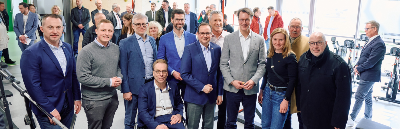 Hendrik Wüst umgeben von mehreren Personen, davon einer im Rollstuhl, in einer Halle mit Fenstern im Hintergrund