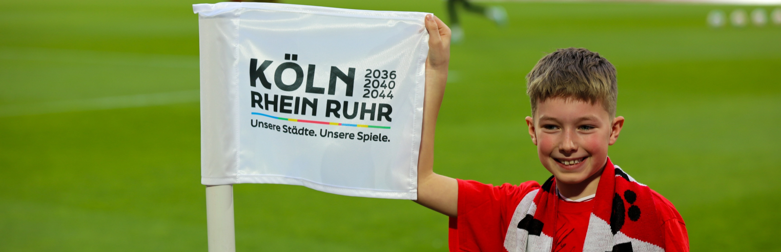 Junge in rotem Trikot auf dem Rasen im RheinEnergiestadion hält die Fahne "KölnRheinRuhr"