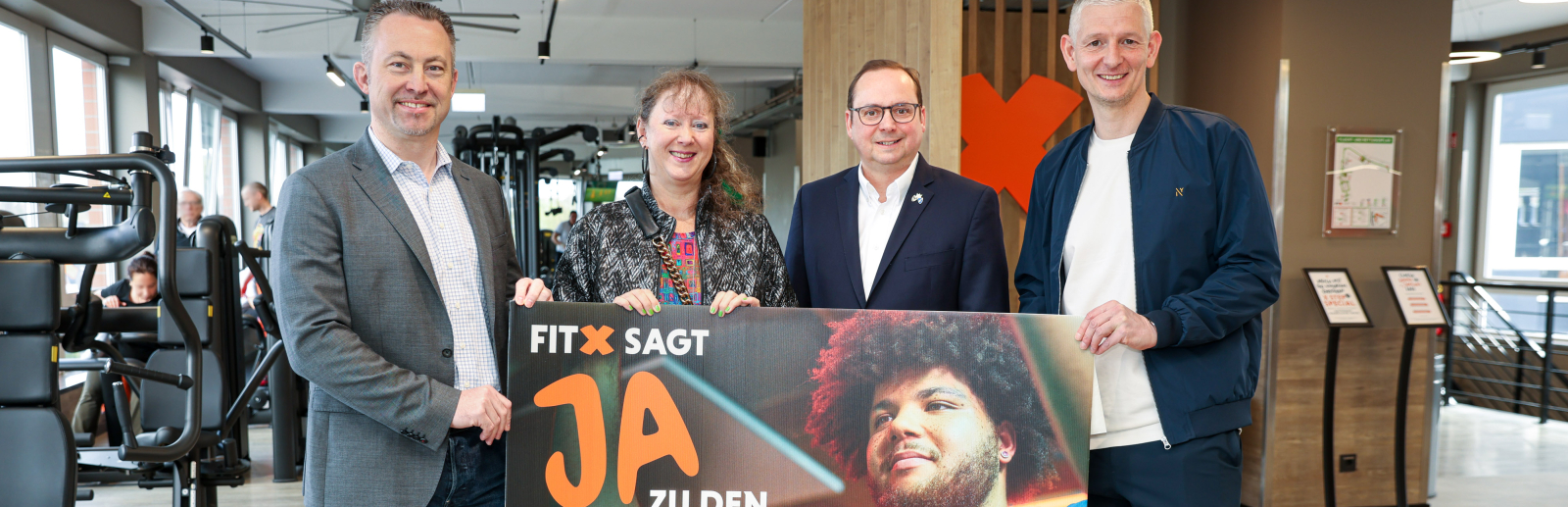 Andrea Milz mit drei Männern, im Hintergrund ist ein Fitnessstudio zu sehen, sie halten ein Banner "Fitx sagt Ja zu Spielen an Rhein & Ruhr"