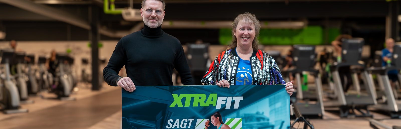 Andrea Milz neben einem Mann in einem Fitnessstudio, beide halten ein Banner zur KRR-Kampagne