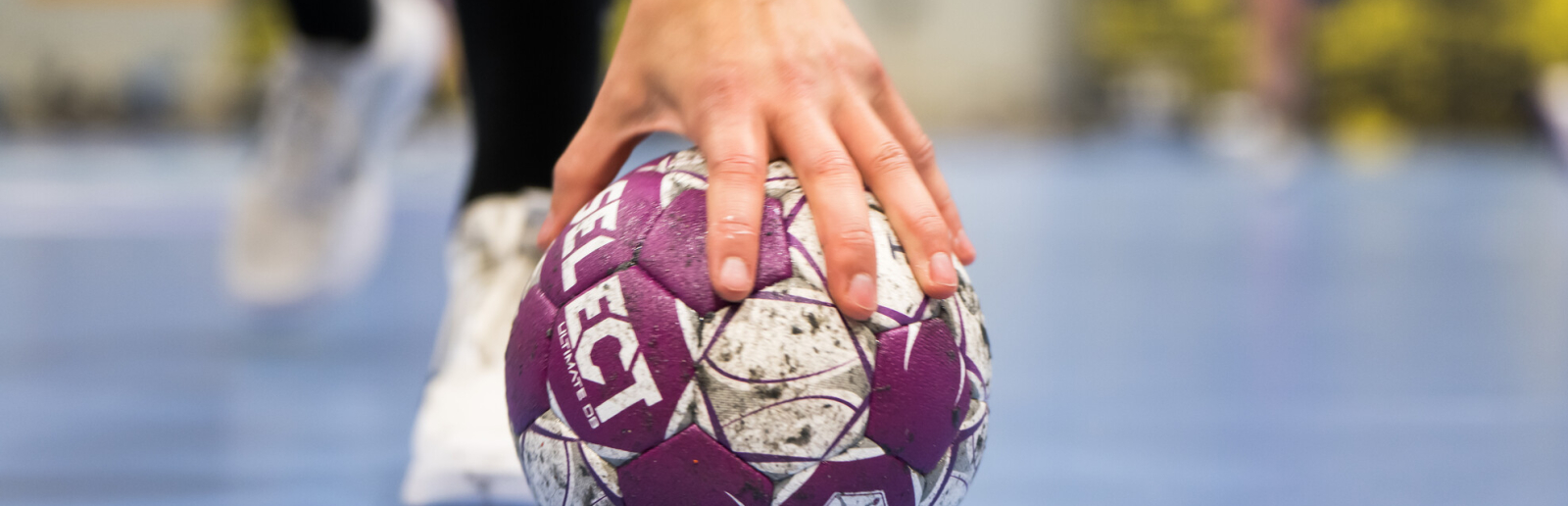 Handball auf einem blauen Boden, im Hintergrund sind Hände zu sehen